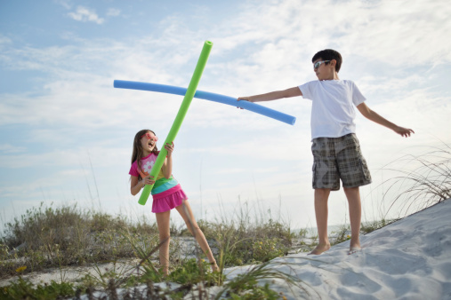 Little children fighting with pool noodles on beach against cloudy sky