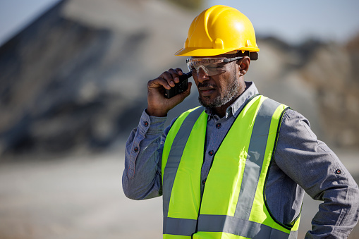 Portrait of industry worker or engineer with hardhat and protective eyewear using walkie-talkie at construction site