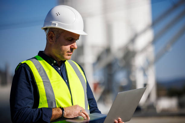 portret poważnego inżyniera w kasku używającego laptopa podczas pracy w zakładzie przemysłowym - fuel storage tank industrial building construction development zdjęcia i obrazy z banku zdjęć