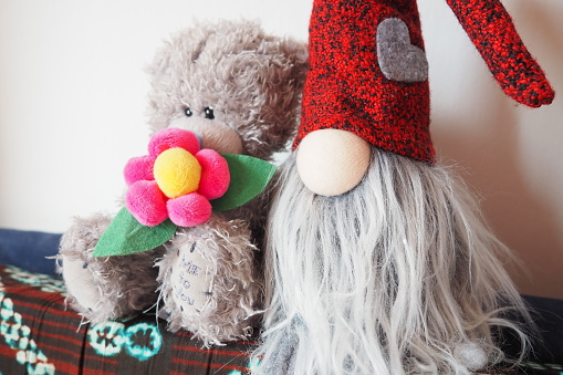 A New Year's gnome in a red hat and a Teddy bear with a flower in his paws sit against the background of a white wall. New Year's mood, the spirit of Christmas. Children's European new fashion toys