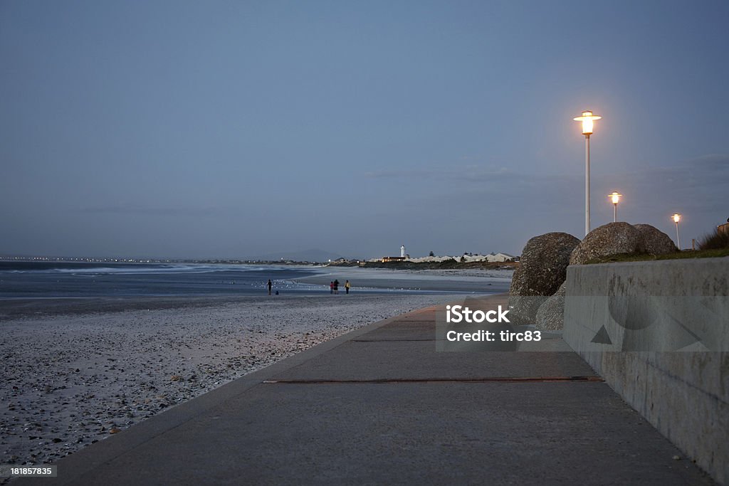 Boardwalk of Cape Town beach at sunset Concrete boardwalk of Lagoon Beach in Cape Town at sunset. Milnerton lighthouse and anonymous dog walkers are pictured in the background Boardwalk Stock Photo