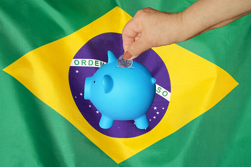 Hand of elderly woman putting coin into piggy bank on Brazil flag background. Hand putting coin to piggy bank. Retirement savings, piggy bank. Concept saving money and retirement fund in Brazil