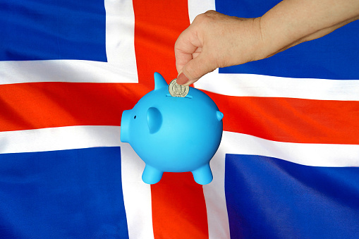 Hand of elderly woman putting coin into piggy bank on Iceland flag background. Hand putting coin to piggy bank. Retirement savings, piggy bank. Concept saving money and retirement fund in Iceland