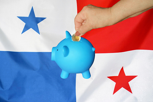 Hand of elderly woman putting coin into piggy bank on Panama flag background. Hand putting coin to piggy bank. Retirement savings, piggy bank. Concept saving money and retirement fund in Panama