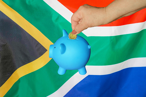Hand of elderly woman putting coin into piggy bank on South Africa flag background. Hand putting coin to piggy bank. Retirement savings. Concept saving money and retirement fund in South Africa