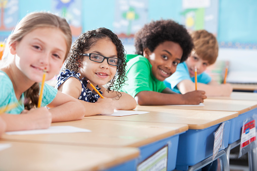 Multi-ethnic elementary school children writing in classroom.  Focus on Hispanic girl wearing eyeglasses (8-9 years).