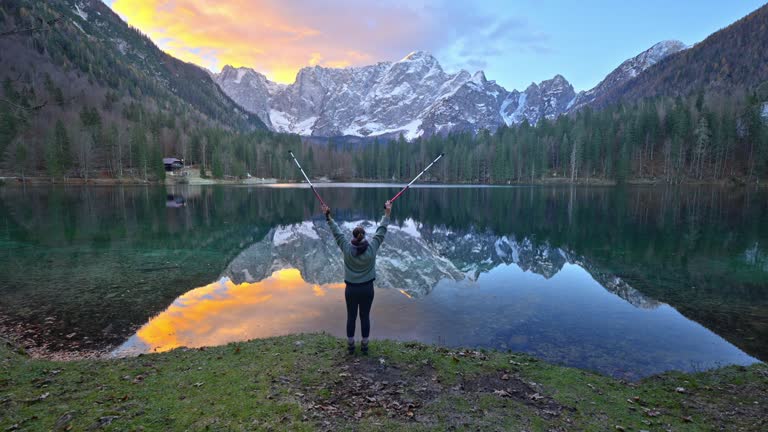 SLO MO Rear View of Woman With Hiking Poles Raising Arms Near Beautiful Fusine Lakes Against Majestic Rocky Mountains at Sunset