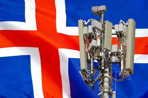 Telecommunications tower with a 5G cellular network antenna agains flag of Iceland. Telecommunication tower of 5G cellular communication. 5G technology usage on telecommunications towers in Iceland