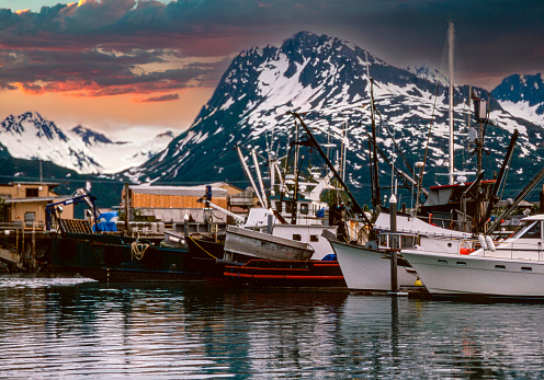 Sunset at an Alaskan harbor captures the stillness of the water and the end of the day for the fishing trade. Snow capped mountains add to the extraordinary scene.