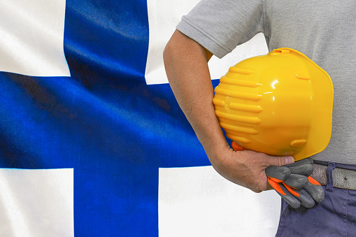 Close-up of hard hat holding by construction worker on Finland flag background. Hand of worker with yellow hard hat and gloves. Concept of Industry, construction and industrial workers in Finland