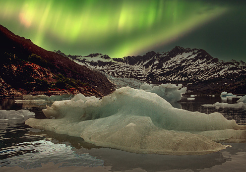 Dramatic lighting  in the sky from the aurora borealis is captured along the rocky coastline of Alaska.