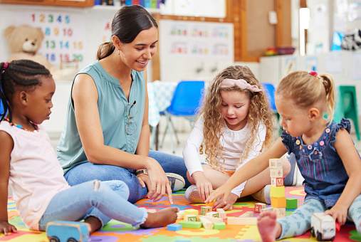 A cute little girl raises her hand to ask or answer a question while sitting at the table with other students and a teacher.