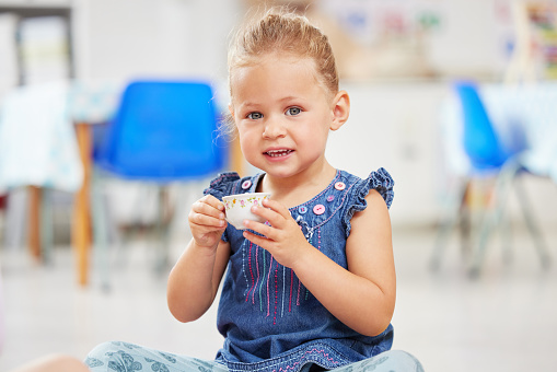 Portrat of one adorable little caucasian girl sitting alone in preschool and holding a small teacup. Smiling child playing and having a tea party at school. Social skills develop children at creche