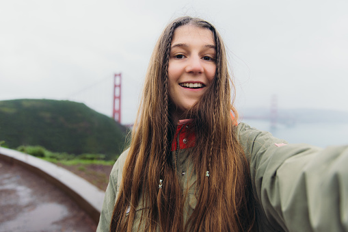 Self-portrait of smiling female influencer with long hair walking at the observation point of Golden Gate bridge during rainy spring calmness morning making selfie and doing video call in San Francisco, the United States