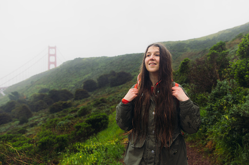 Portrait of smiling female traveller with long hair and backpack walking on the fresh green hill with view of big red bridge during rainy spring calmness morning in San Francisco, the United States