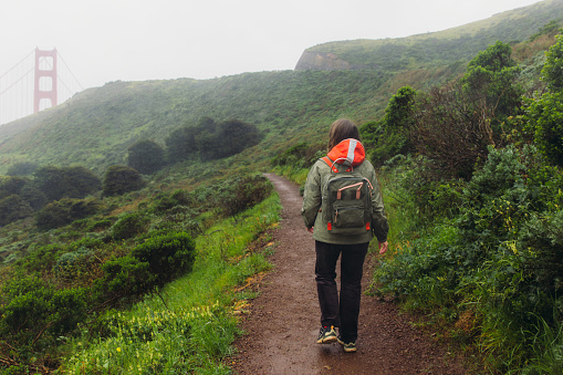 Rear view of female traveller with long hair and backpack walking on the fresh green hill with view of big red bridge during rainy spring calmness morning in San Francisco, the United States