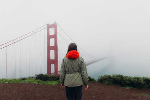 Rear View of female traveller with long hair walking at the observation point of big red bridge during rainy spring calmness morning in San Francisco, the United States