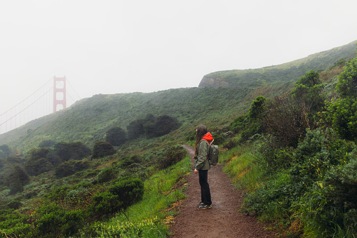Side View of female traveller with long hair and backpack walking on the fresh green hill with view of big red bridge during rainy spring calmness morning in San Francisco, the United States