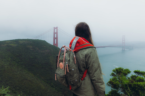Rear View of female traveller with long hair walking at the observation point of big red bridge during rainy spring calmness morning in San Francisco, the United States