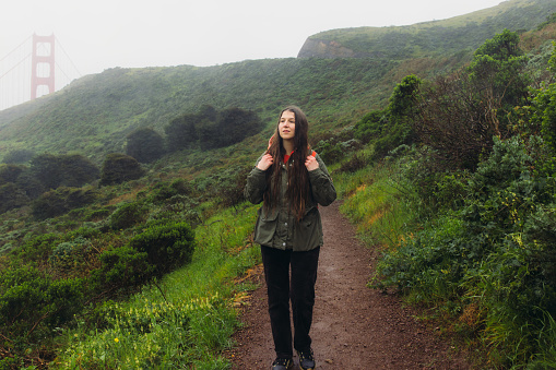 Front View of female traveller with long hair and backpack walking on the fresh green hill with view of big red bridge during rainy spring calmness morning in San Francisco, the United States