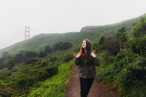 Front View of smiling female traveller with long hair and backpack walking on the fresh green hill with view of big red bridge during rainy spring calmness morning in San Francisco, the United States