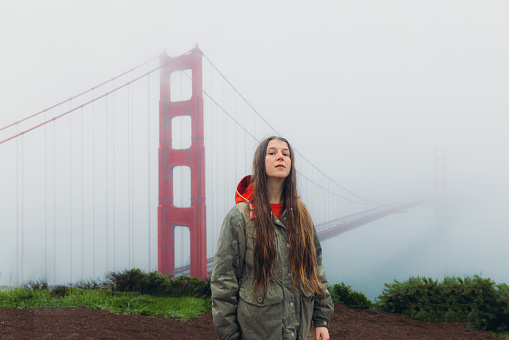 Portrait of female traveller with long hair walking at the observation point of big red bridge during rainy spring calmness morning in San Francisco, the United States