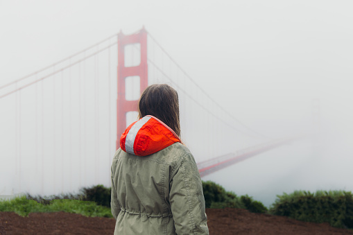 Rear View of female traveller with long hair walking at the observation point of big red bridge during rainy spring calmness morning in San Francisco, the United States