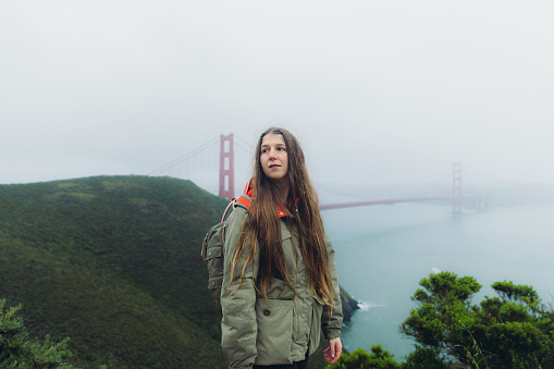 Portrait of female traveller with long hair walking at the observation point of big red bridge during rainy spring calmness morning in San Francisco, the United States
