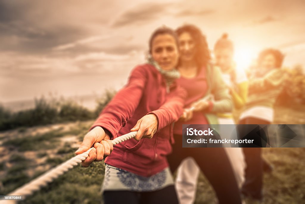 Group of people engaging in tug of war Women using teamwork to pull a rope during a tug of war.  Four women are pulling on the rope. The rope extends off the left side of the image.  The team the women are pulling against is not visible.  The hands of the woman in front are in sharp focus.  The focus becomes more blurred as the eye travels farther down the rope.  The women are bracing with their legs and their faces are strained.  There is grass under their feet and a gray sky overhead.  The sun is low in the sky behind them. Tug-of-war Stock Photo