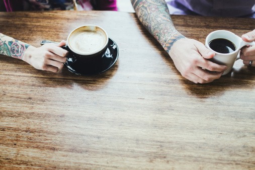 A trendy hipster man and woman with full arm sleeve tattoos smile and talk as they enjoy a coffee and latte at a bright cafe, sitting at a large bamboo wood table.  Hoprizontal overhead shot with copy space.