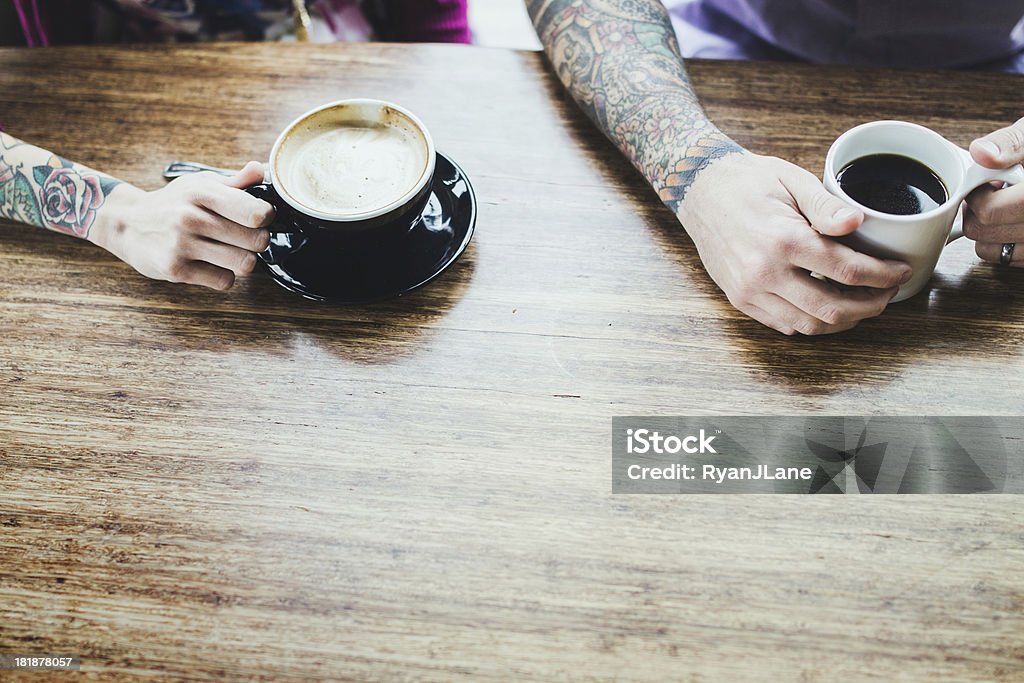 Modern Couple At Coffee Shop A trendy hipster man and woman with full arm sleeve tattoos smile and talk as they enjoy a coffee and latte at a bright cafe, sitting at a large bamboo wood table.  Hoprizontal overhead shot with copy space. Tattoo Stock Photo