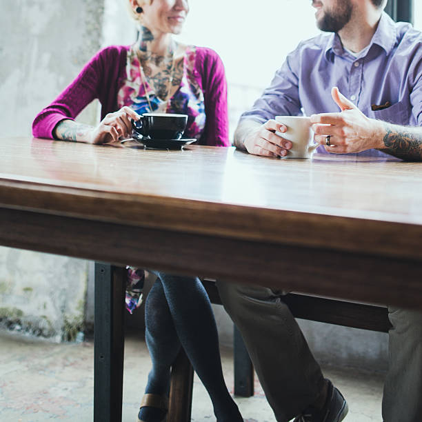 The hands of two young woman lifting their cups of coffee, cappuccinos. They are sitting at a round table in a cafe or coffee shop, talking and gesturing.