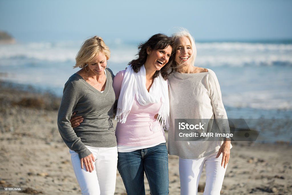 Trio of women walking at the beach Three smiling mature women embracing one another on a rocky, seaweed covered sandy beach.  The ocean and clear blue skies are behind them. Mature Women Stock Photo