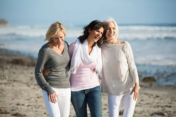 Three smiling mature women embracing one another on a rocky, seaweed covered sandy beach.  The ocean and clear blue skies are behind them.