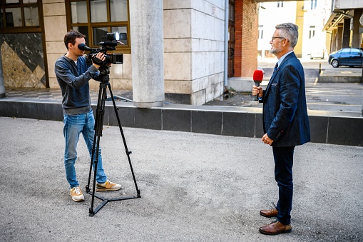 A male television reporter holding a microphone, is standing outdoors in front of a building. A male camera operator is filming him.