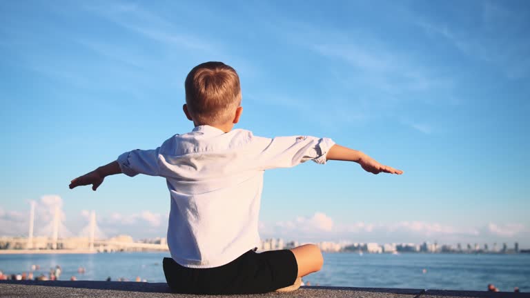 Child boy sit on embankment by water in center of city of St. Petersburg, spreading and shaking arms to sides in form of airplane in sense freedom. Tourist look sights and admires summer landscape.