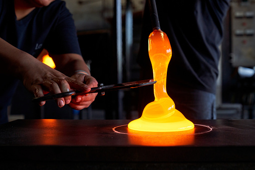 Crop male glassmiths in uniform in process of forming hookah vase using blowpipe and sheers while working at professional glassblowing factory together