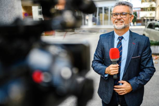 A male television reporter holding a microphone, is standing outdoors in front of a building. A male camera operator is filming him.