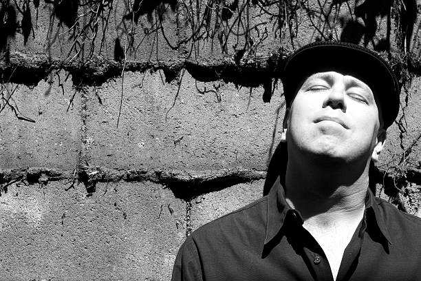 Man and Brick Wall Close-Up #2 of 2 "Black & White photo of a man in a hat, standing, relaxed, up against a brick wall, head back, basking in the bright sunlight. High contrast, bright light and rich shadows from the thick mortar squeezed between the bricks form a rich tapestry of light and dark. Bright levels deliberately over-enhanced to reinforce the sunlight vs. shadow contrasts." 2004 2004 stock pictures, royalty-free photos & images