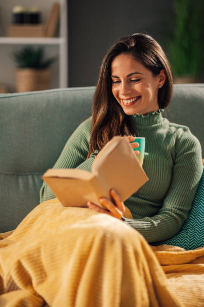 istock Smiling woman relaxing on a couch at home covered in blankets reading. 1818907825