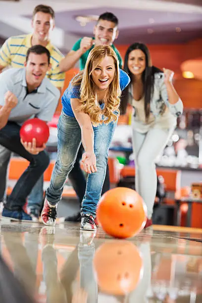 Group of friends cheering in the background while the girl is throwing a bowling ball.
