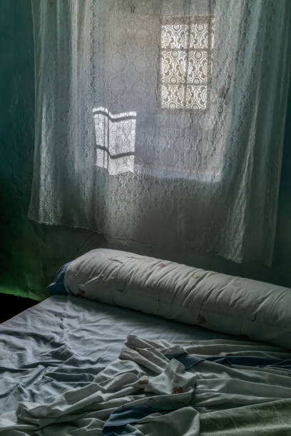 Castilla y León, Spain. Old village room with a small window draped in curtains and a unmade bed.
An ancient village room exudes a charm of yesteryear and a picturesque atmosphere. The window, adorned with delicate curtains, allows soft natural light to filter in. The worn walls tell tales of days gone by. In the center, an unmade bed with sheets slightly disheveled suggests a moment frozen in rural tranquility.
