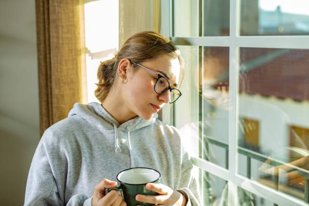 Sorrowful young woman with glasses looking through the window