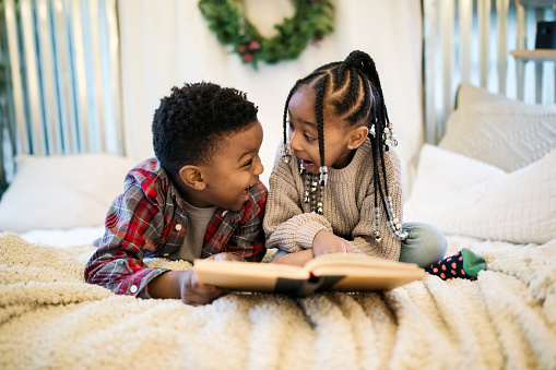 A cute African American brother and sister read a book together on their family vacation for the Christmas holiday, a wreath hanging on the wall behind them.
