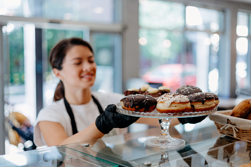 In this shot, the mid-adult woman holds a plate with precision, presenting a selection of donuts that reflect her artistry in both flavor and aesthetics, inviting customers to savor the delectable creations