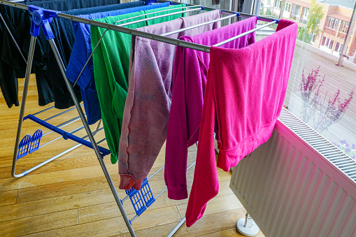 Colorful clean clothes hanging on collapsible drying rack next to a white heating radiator, indoor excessive humidity problem