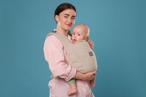 Mother holding her child in sling (baby carrier) on light blue background