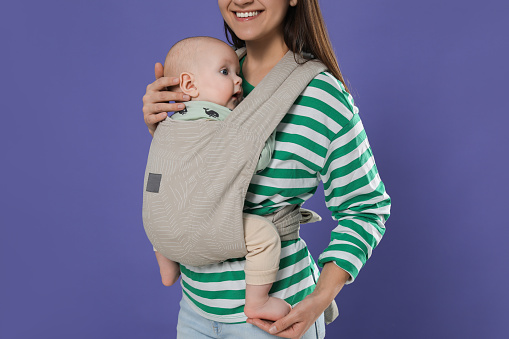 Mother holding her child in sling (baby carrier) on purple background, closeup