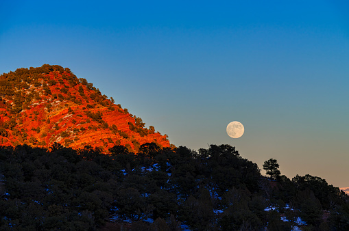 Moonrise Over Red Rock Canyon and Mountains - Scenic view with full moon, blue sky and orange/red rock warmly lit at sunset. Moon in lower third of sky with copy space.