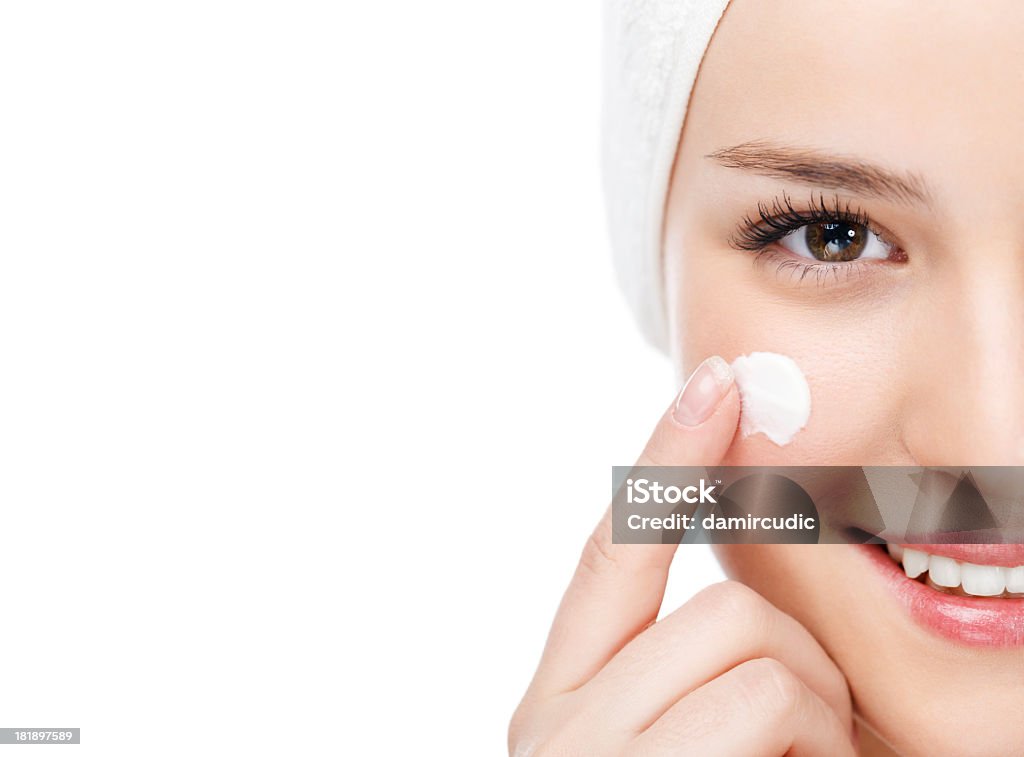 Woman applying moisturizer This is a close-up portrait of a young and beautiful, smiling woman with brown eyes,who is applying a mousturizer on her face. The woman has a white towel on her head. The women is isloated on white background.  Moisturizer Stock Photo